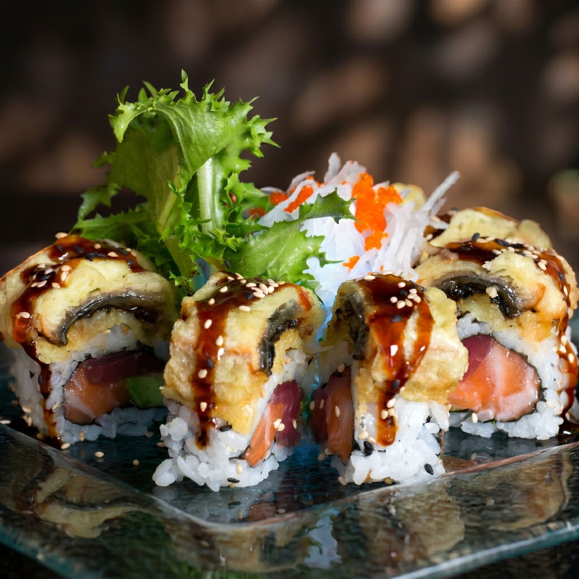 Sushi rolls topped with sauce on a glass plate, garnished with greens and shredded carrot