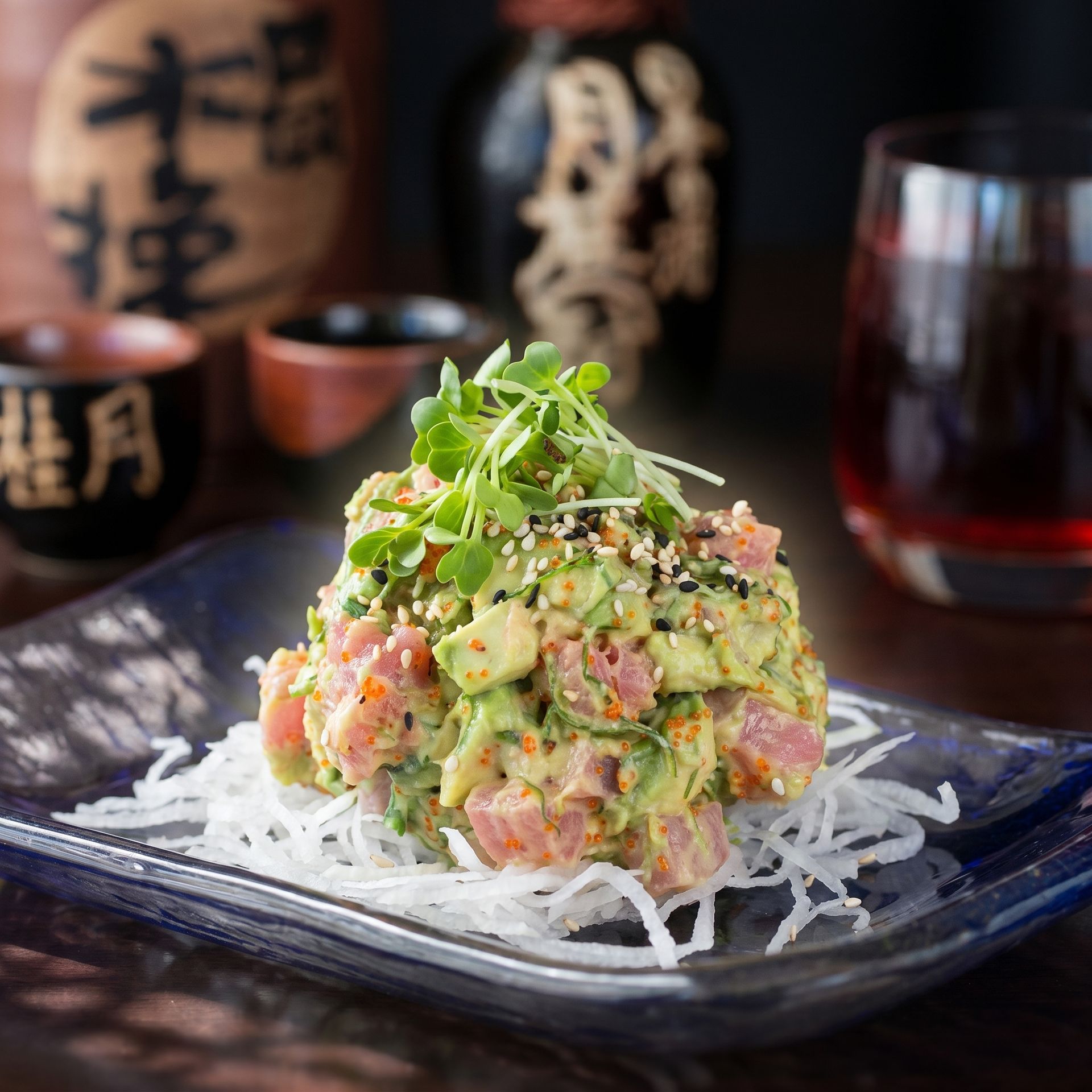 Tuna salad mound on a dark plate, topped with greens, with sake cups and bottles in the background