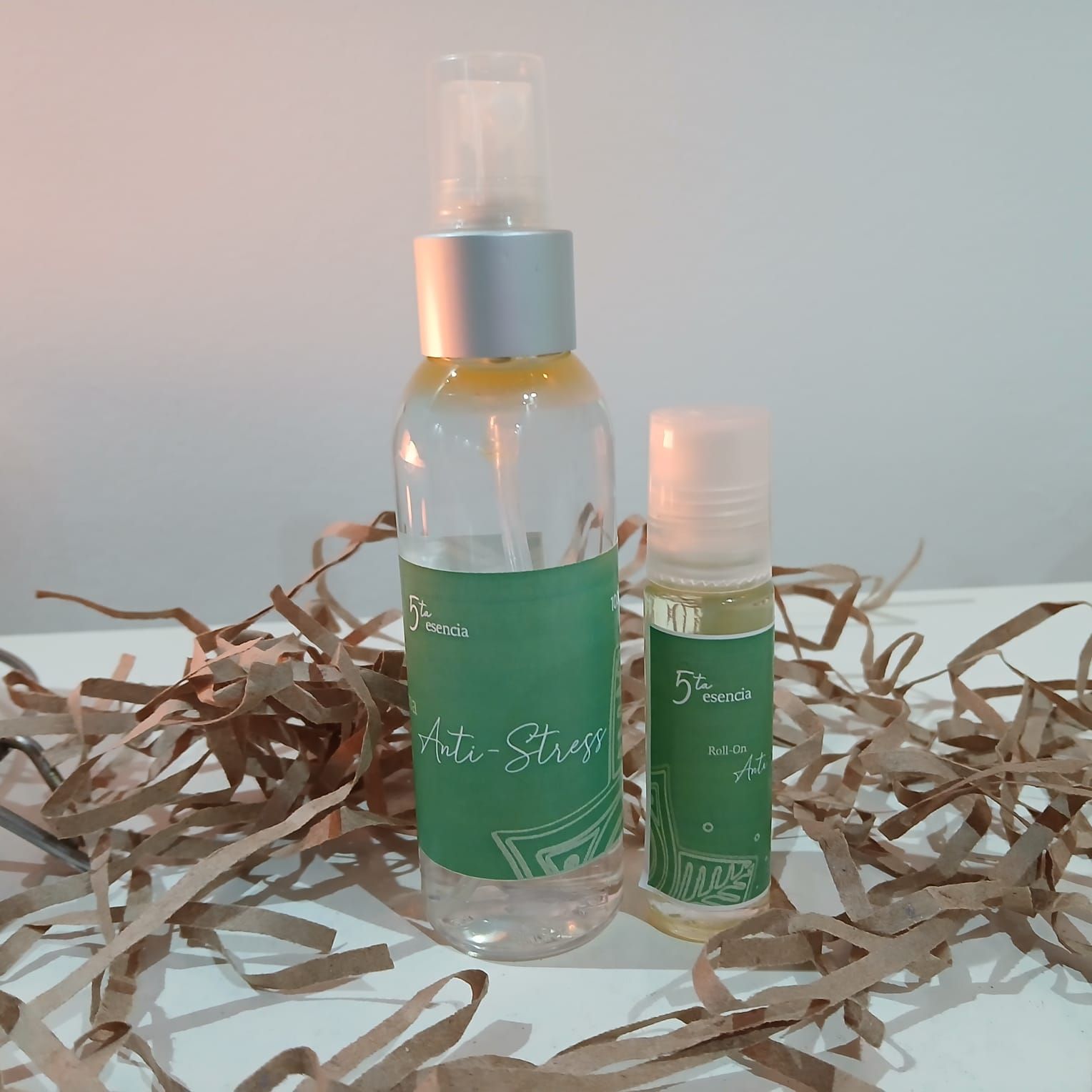 Frasco de spray y roll-on con aroma a verde, relleno de papel triturado y tapones plateados sobre un fondo pálido.
