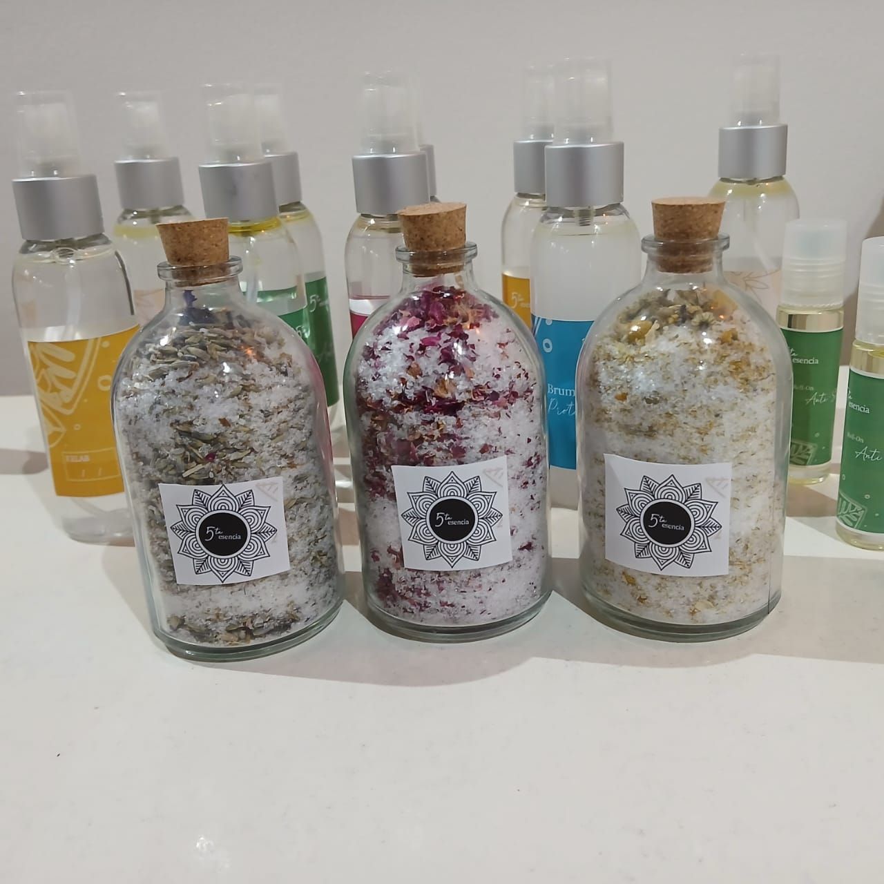 Tres botellas decorativas brillantes con tapón de corcho sobre una mesa blanca, delante de botellas de colores.