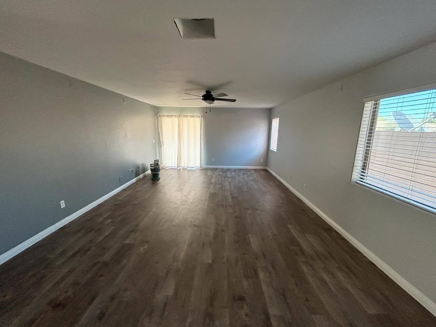 Empty living room with dark wood-look flooring, gray walls, and a sliding glass door. A ceiling fan is centered above.