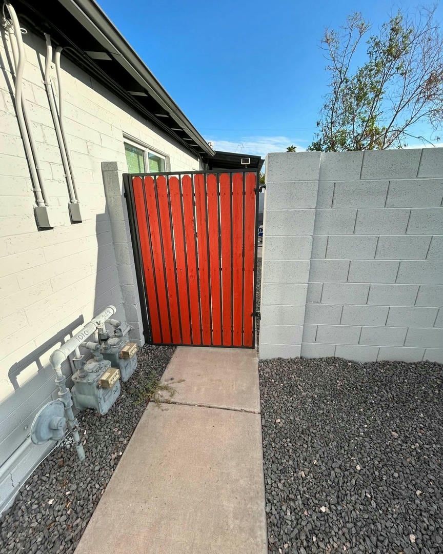 A bright orange wooden gate in a black frame set in a white brick wall with a concrete path.