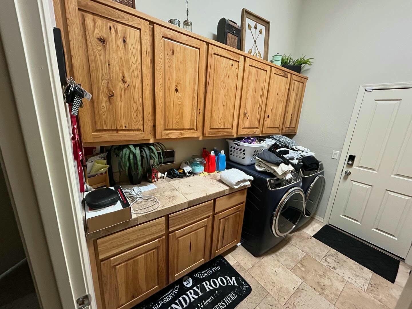 Laundry room with wooden cabinets, black washer and dryer, and a laundry room doormat. A white door is on the right.