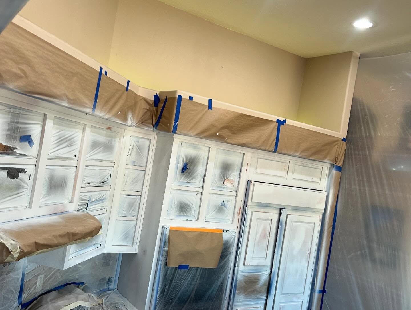 Kitchen cabinets covered in plastic and paper, ready for painting. Tape secures the coverings, with off-white paint visible.