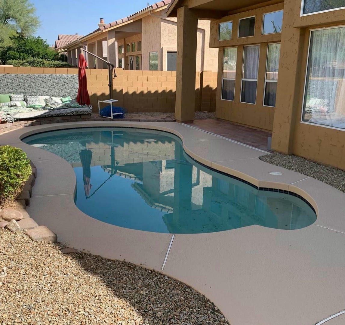 A backyard pool with a light-colored concrete deck. The pool is surrounded by small rocks and a covered patio.