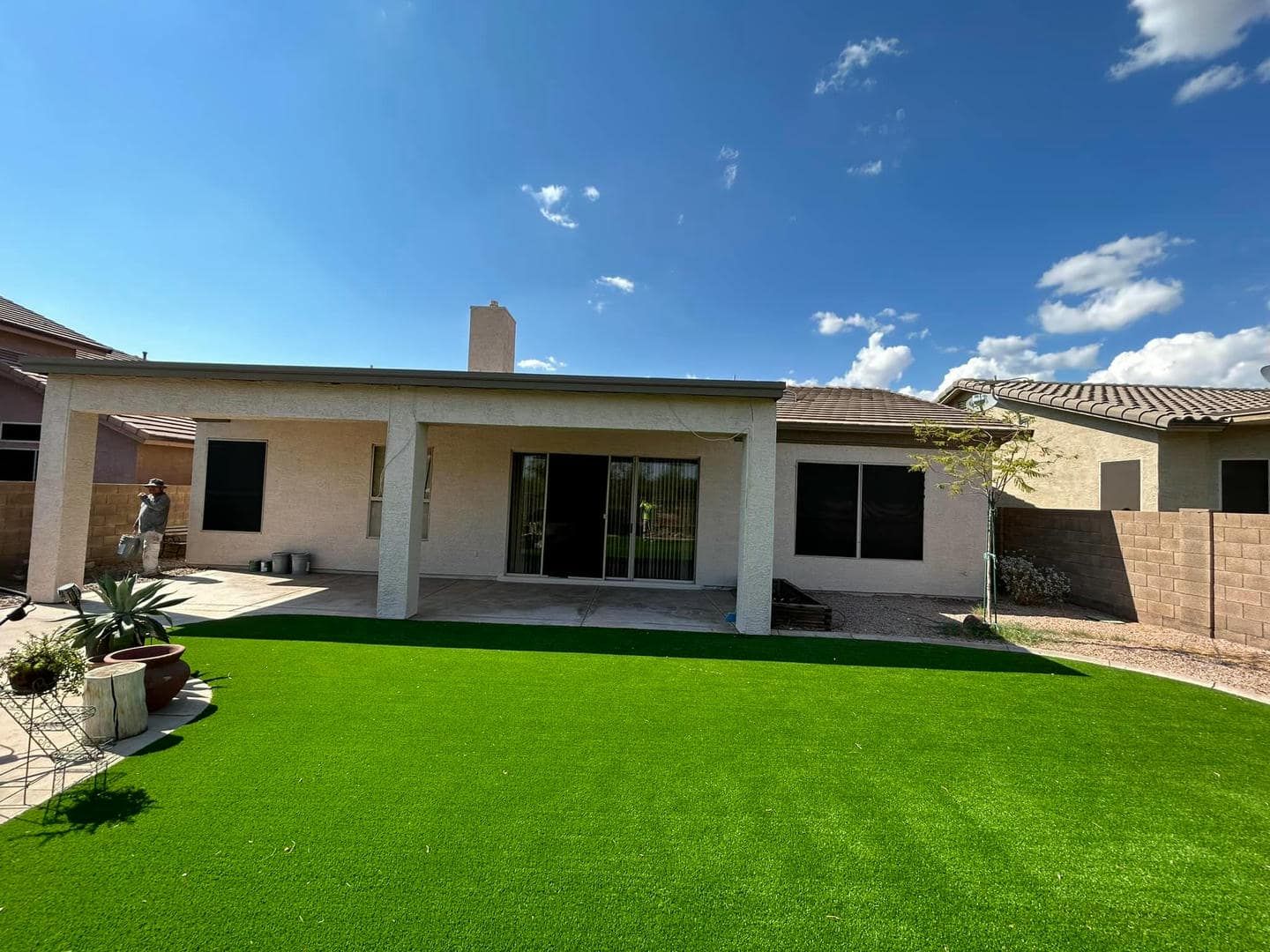 Backyard with green lawn, covered patio, and a clear blue sky. Houses and a brick wall are visible in the background.