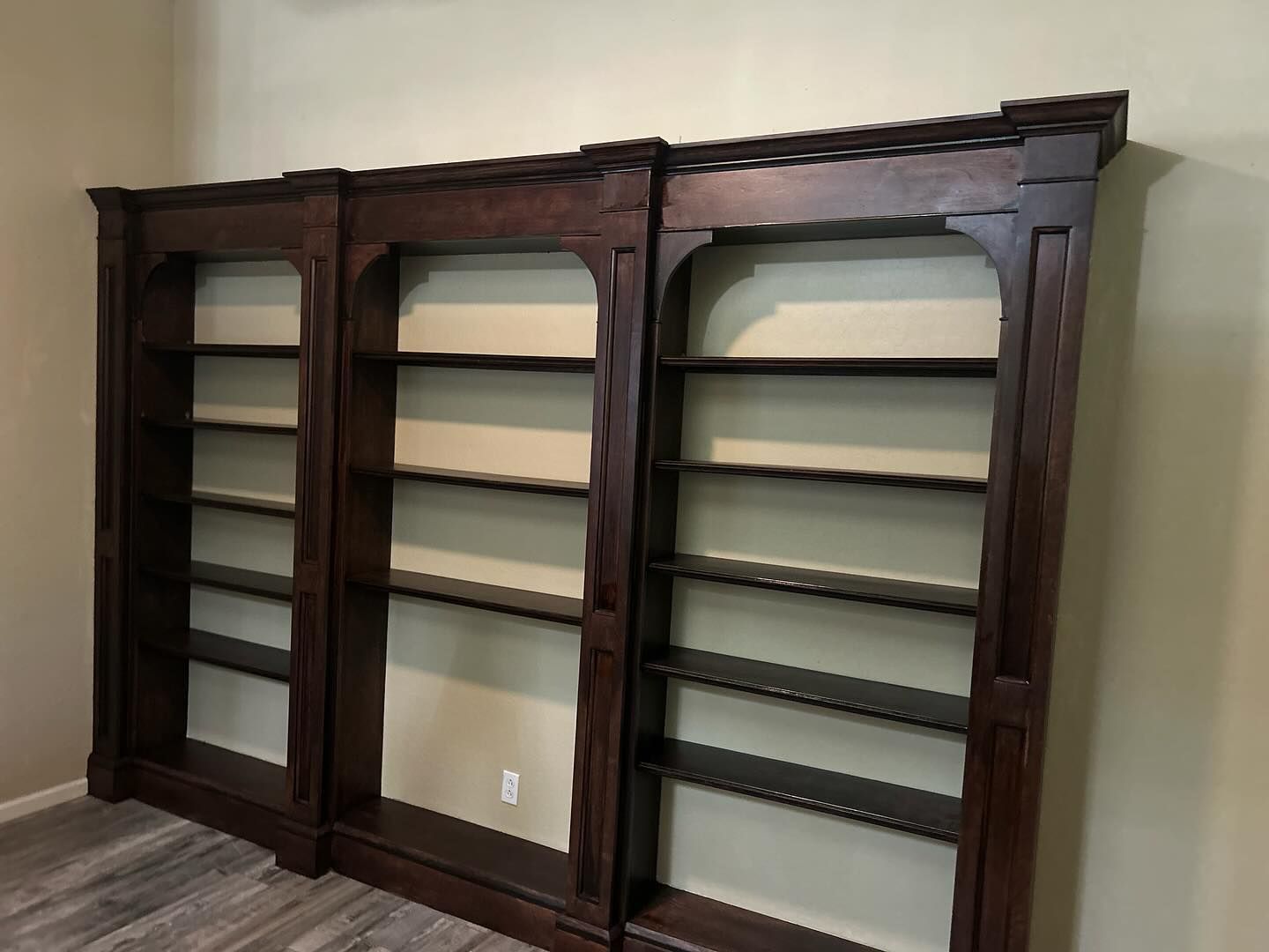 Three dark brown wooden bookshelves against a beige wall. The shelves have arched tops and are empty.