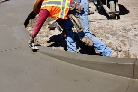 Construction worker smoothing wet concrete curb with a trowel. Wearing safety vest and blue jeans.