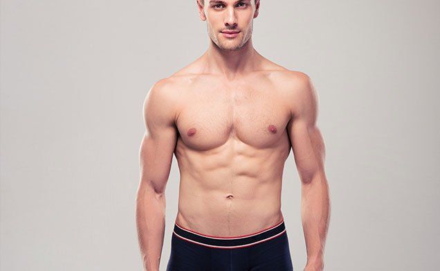 A shirtless man in black underwear is standing in front of a gray background.