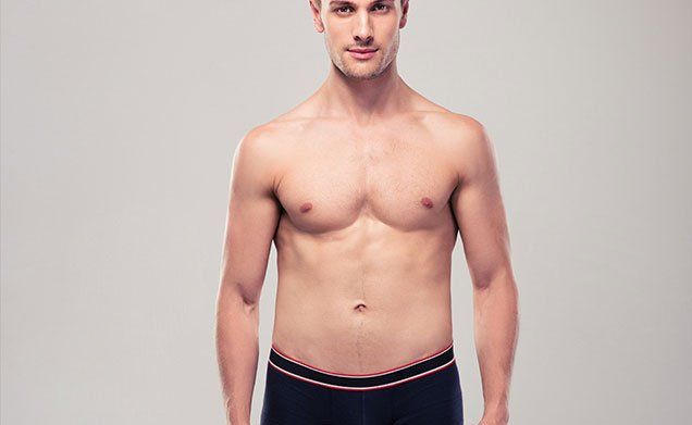 A shirtless man in black underwear is standing in front of a gray background.