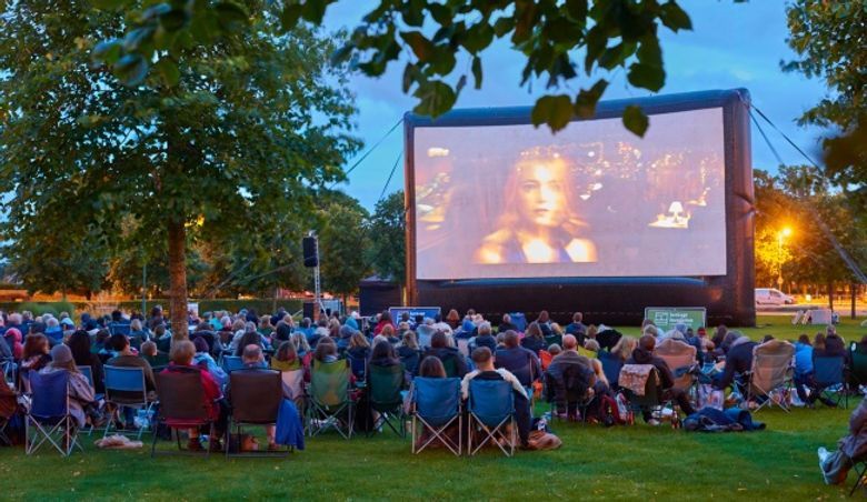 People in lawn chairs watch a movie on a large screen in a park at dusk.