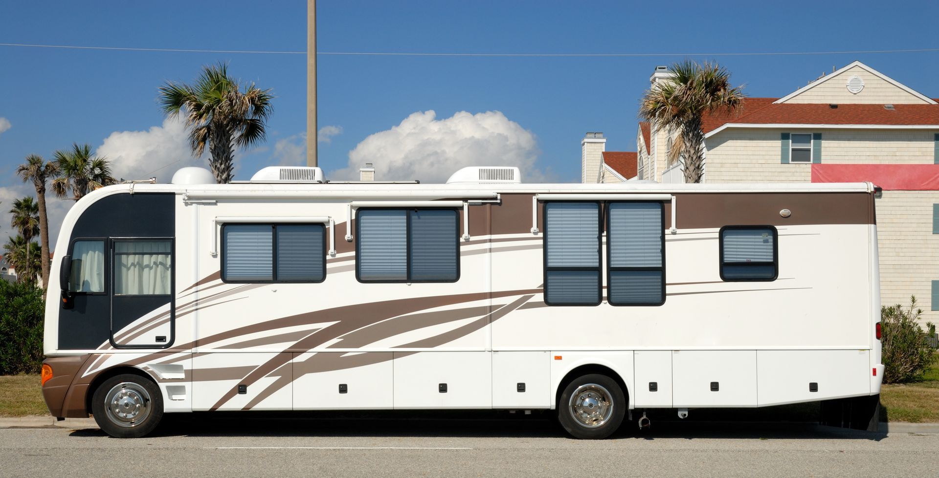 White and brown RV parked on a street near a beach. Palm trees and a building are in the background.