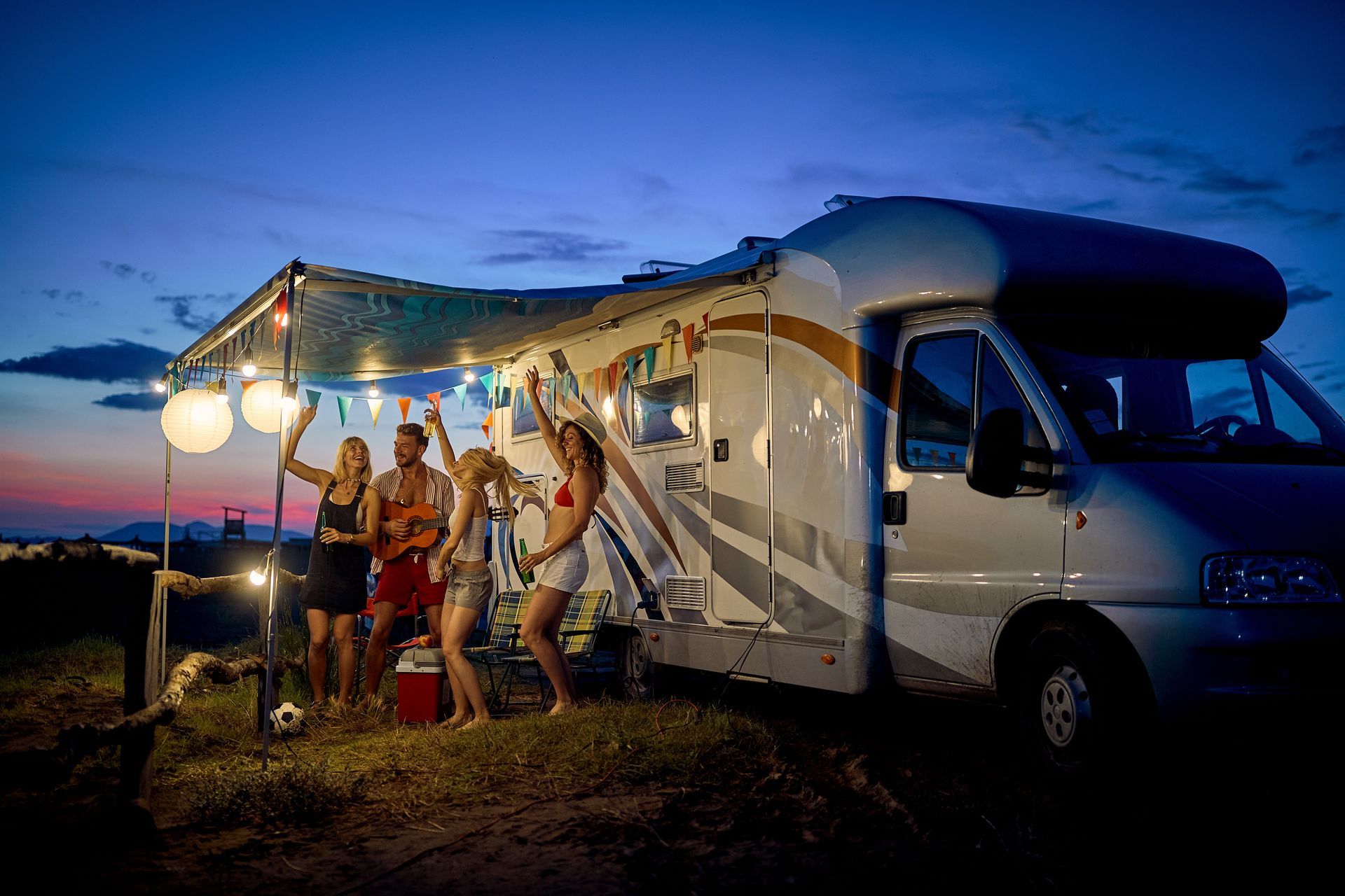 Friends celebrating outdoors at dusk with an RV. Strings of lights, drinks, and music.
