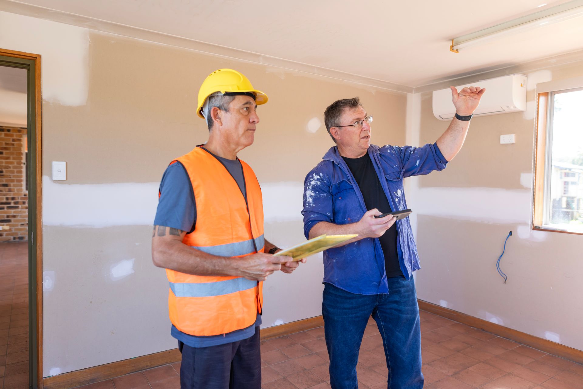Two construction workers are standing in an empty room looking at the ceiling.