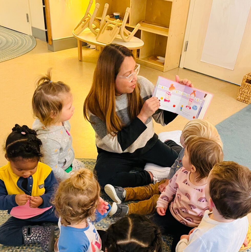 A guide is sitting on the floor reading a book to a group of Montessori children
