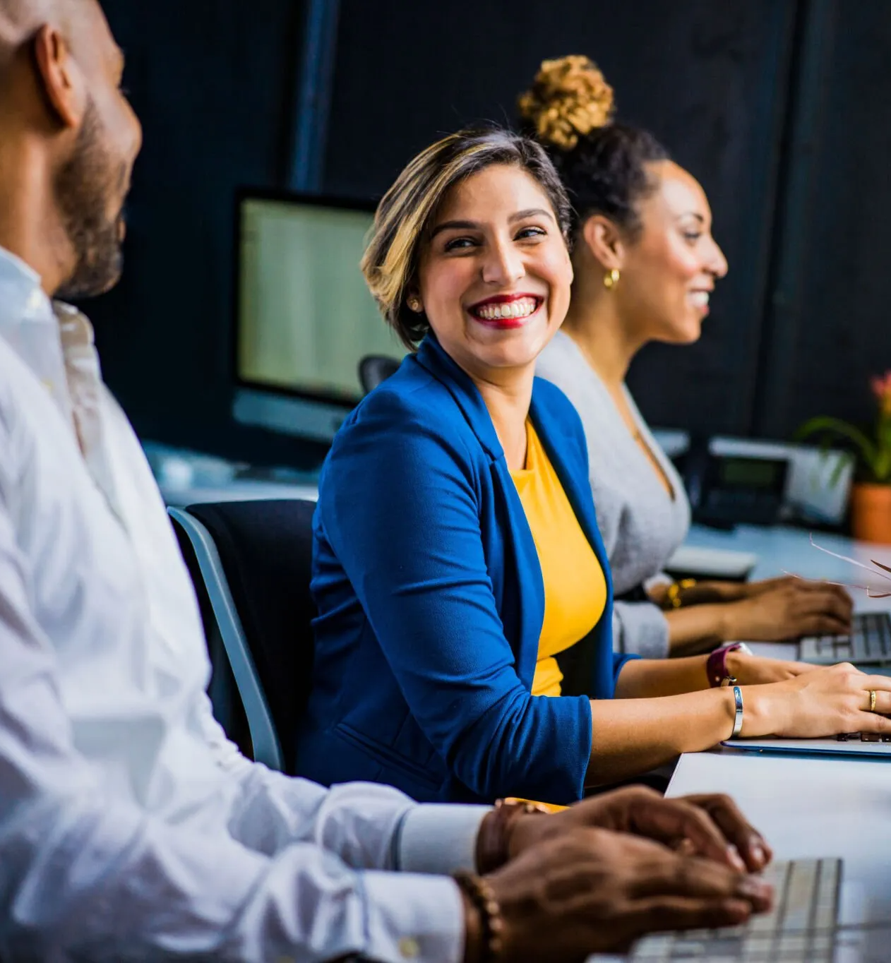 A woman in a blue jacket is smiling while sitting at a desk with other people.