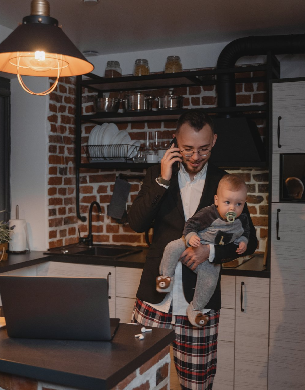 A man is holding a baby and talking on a cell phone in a kitchen.