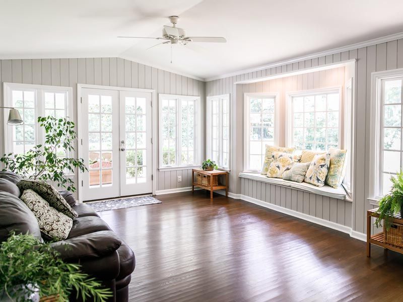 Bright sunroom with large windows, dark wood floors, a black sofa, and a white bench with floral pillows