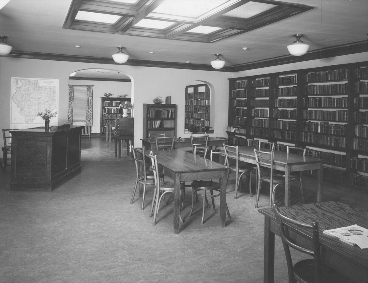 A black-and-white view of a quiet library with wooden tables, chairs, bookshelves lining the wall, and a coffered ceiling.
