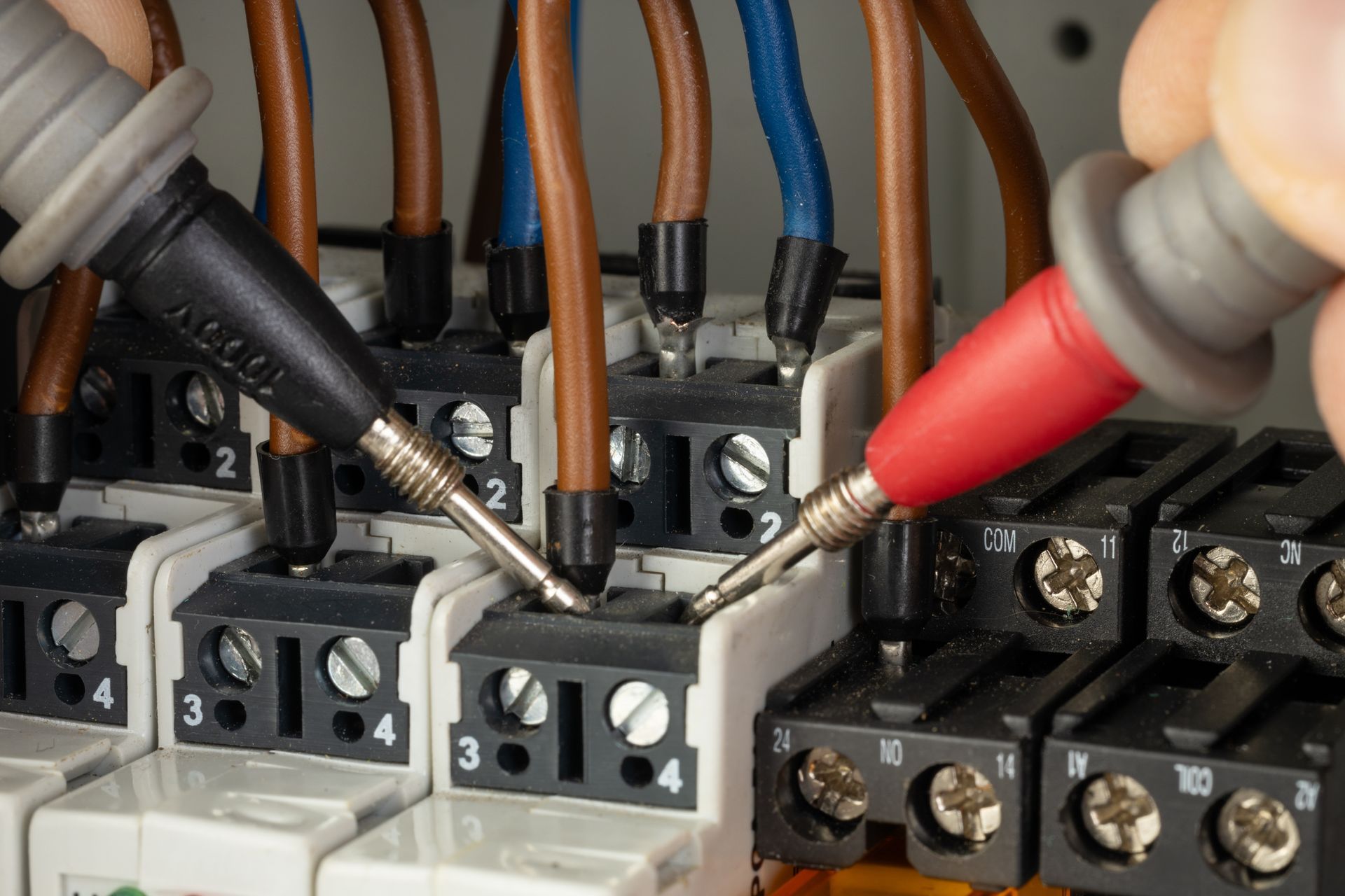 Multimeter probes testing electrical terminals in a circuit breaker panel with brown and black wires.