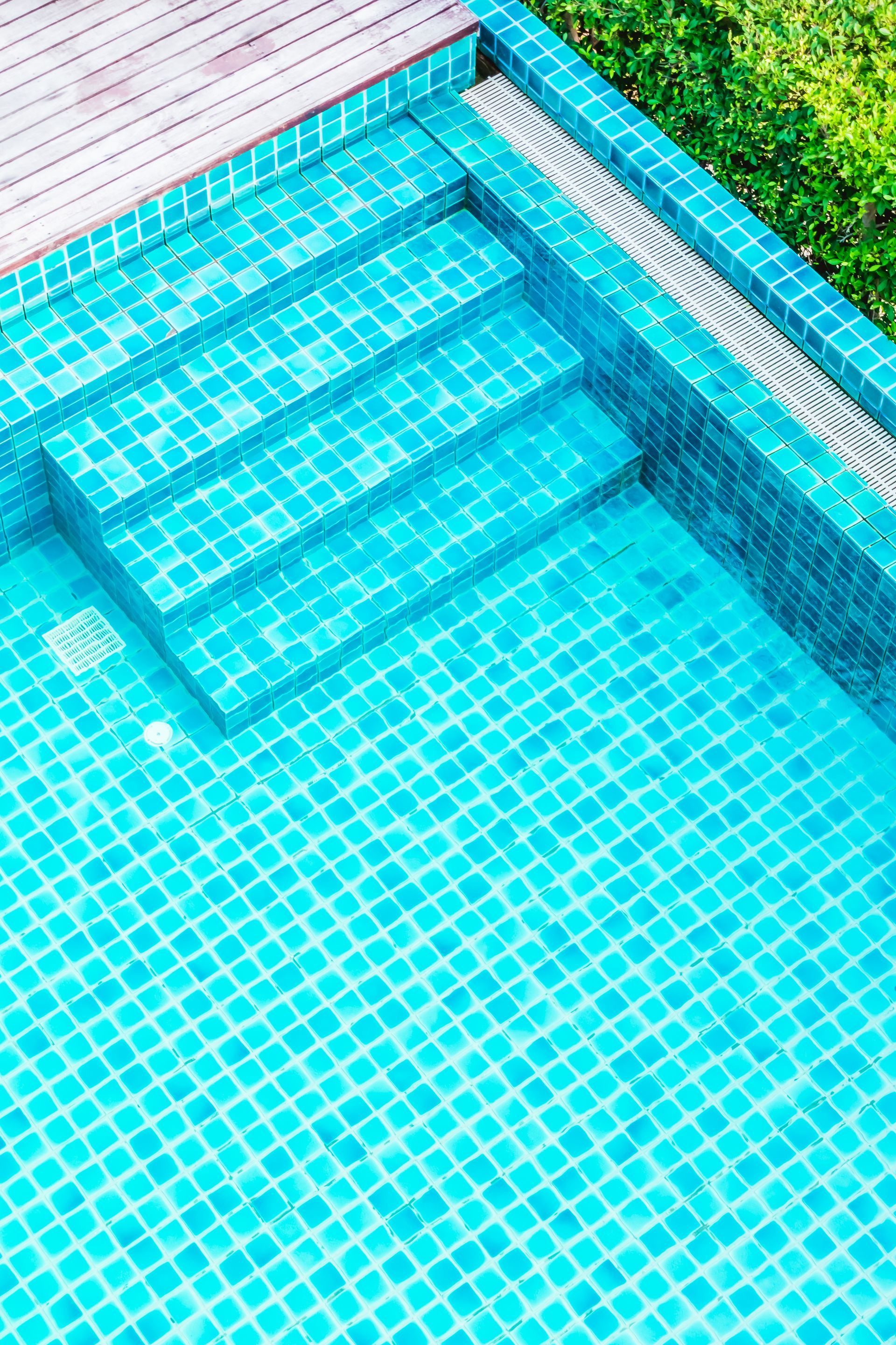A high-angle view of a bright turquoise swimming pool featuring tiled stairs leading into the water next to a wood deck.