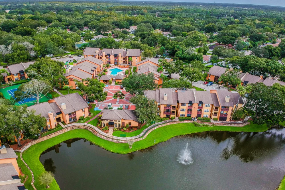 Aerial view of a tan apartment complex with a swimming pool and central courtyard, situated beside a lake with a fountain.