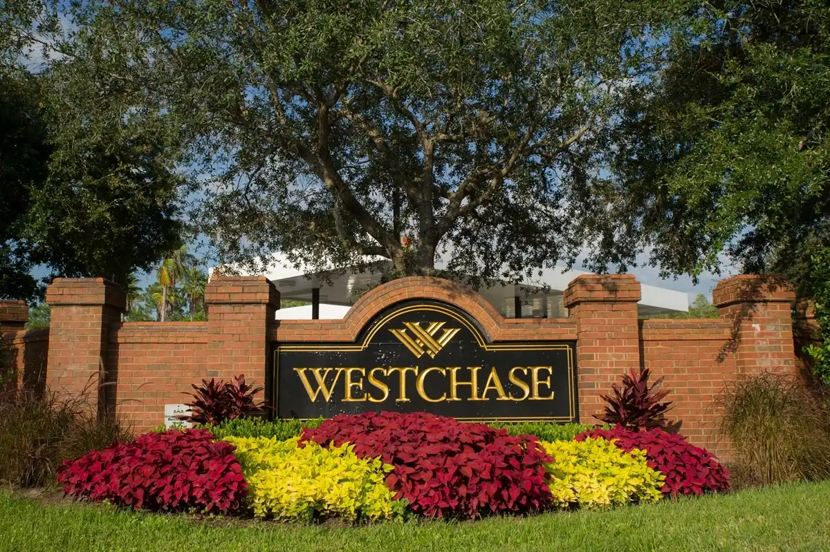 A brick sign for Westchase community, featuring gold lettering, a logo, and a garden bed of red and yellow flowers.