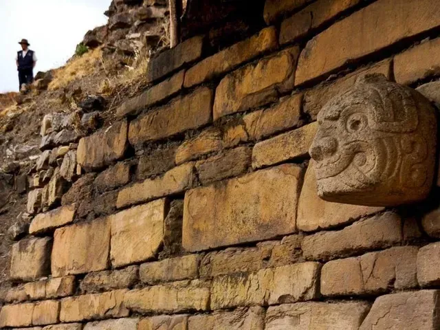 Antiguo muro de piedra con relieve de rostro tallado y una persona de pie en el acantilado rocoso de arriba.