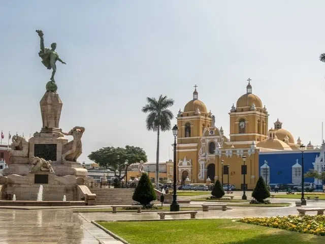 Plaza con una estatua de bronce, palmeras y una catedral de cúpula amarilla junto a un edificio azul.