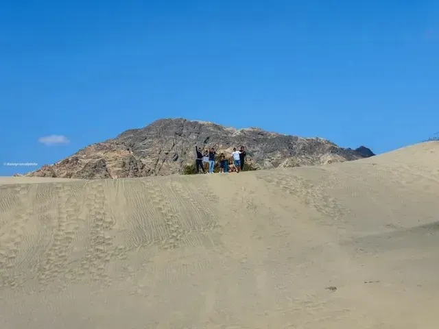 Un pequeño grupo de personas de pie sobre una duna de arena con una colina rocosa bajo un cielo azul despejado.