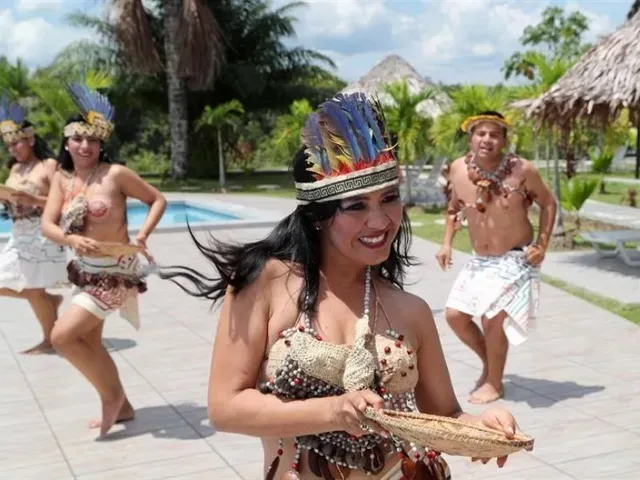 Bailarines con coloridos trajes actúan al aire libre junto a una piscina bajo palmeras.