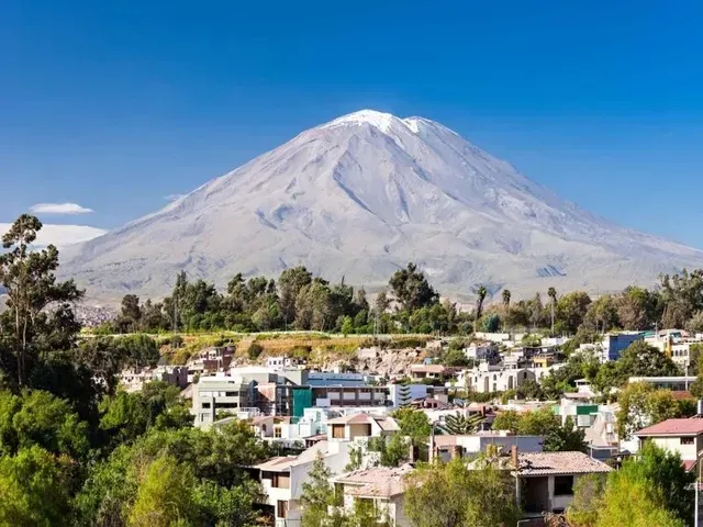 Un volcán cubierto de nieve se alza sobre un pequeño pueblo con árboles verdes y cielo azul.