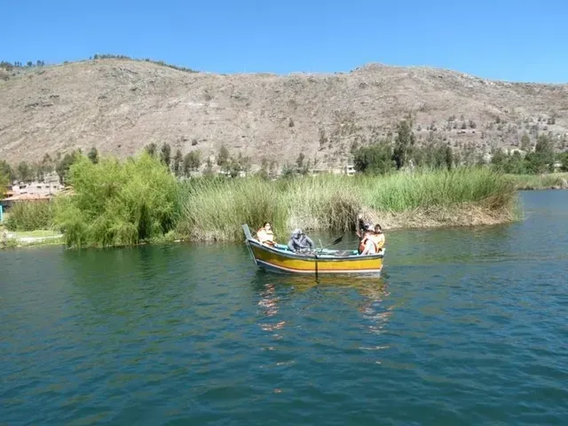 Una pequeña barca con gente en un lago azul tranquilo, cerca de juncos verdes y una ladera rocosa bajo un cielo despejado.