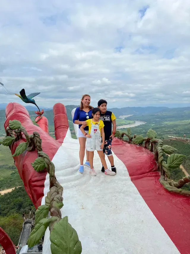 Tres personas posando en un mirador panorámico rojo y blanco con montañas y nubes al fondo.
