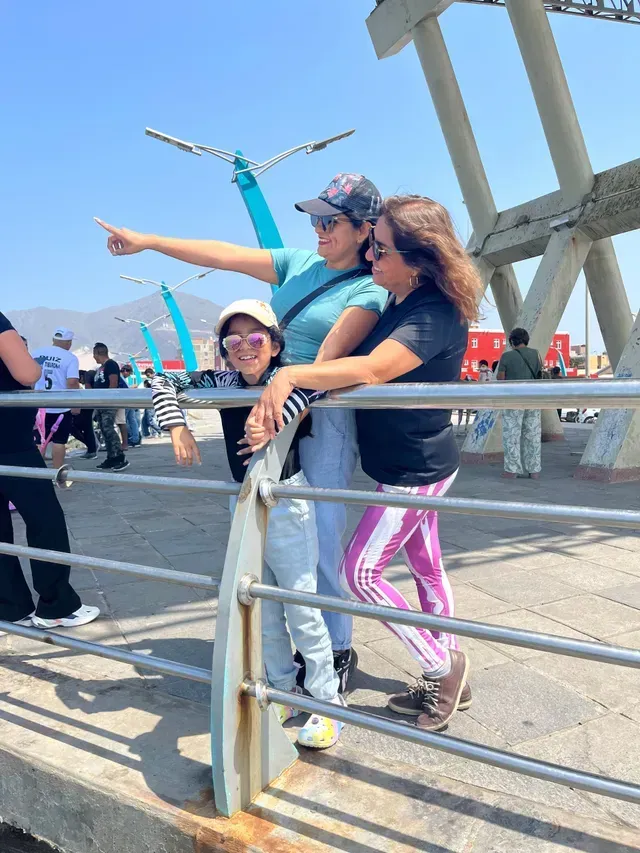 Tres personas posan en un puente soleado, una de ellas señalando hacia adelante, con barandillas de metal