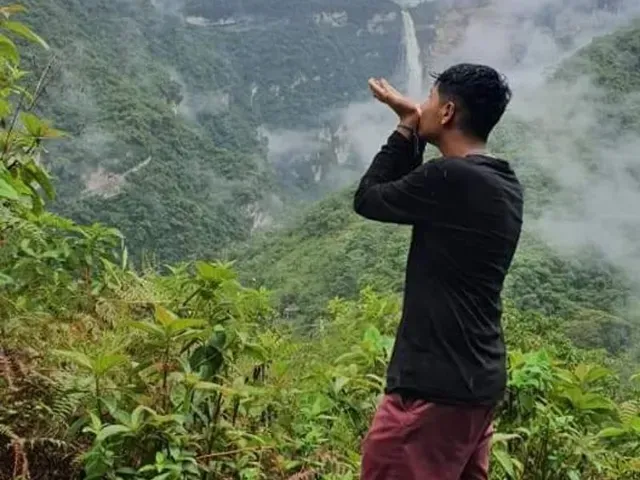 Una persona vestida con camisa negra y pantalones granates se encuentra en frondosas montañas