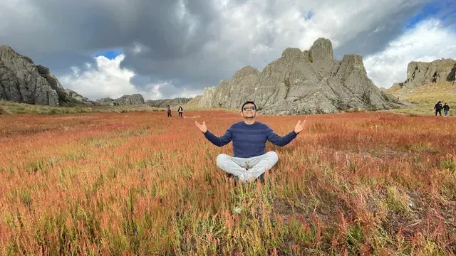 Persona meditando en un prado de flores silvestres con montañas rocosas y un cielo nublado.