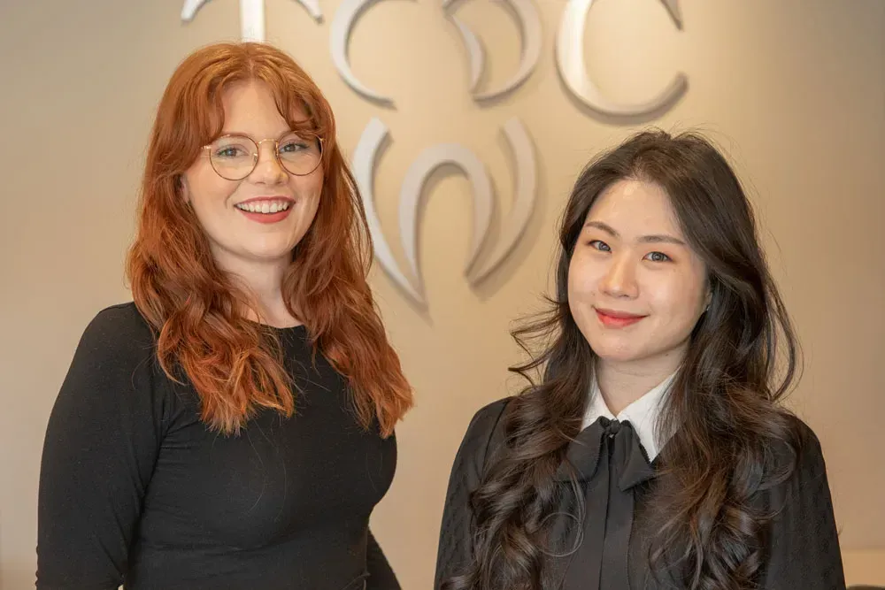 Two women smiling in front of a logo. One with red hair and glasses, the other with dark hair.