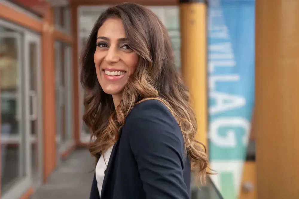 Woman smiles, wearing a navy blazer, outdoors. Background shows a building with a blue and white sign.