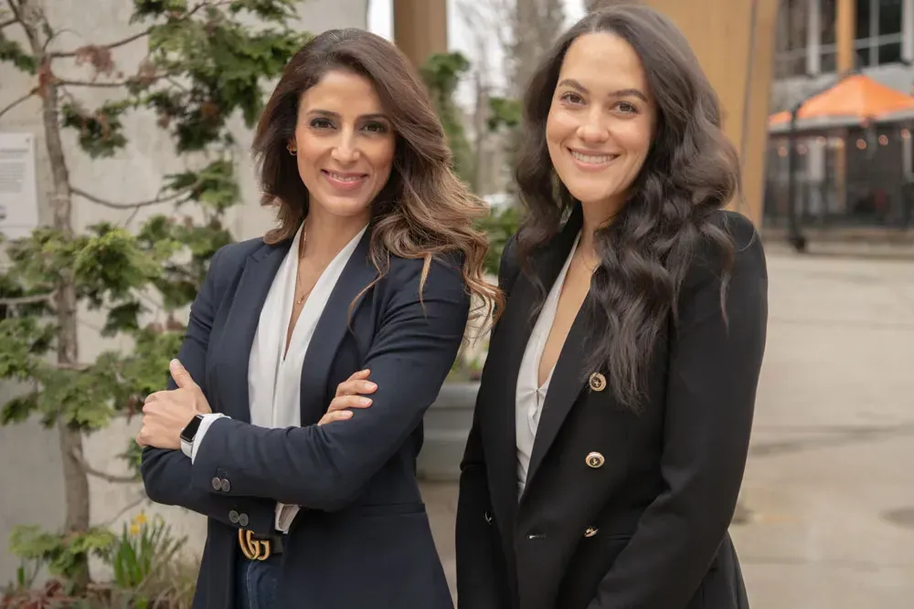 Two women in business suits smiling outdoors. One has arms crossed.