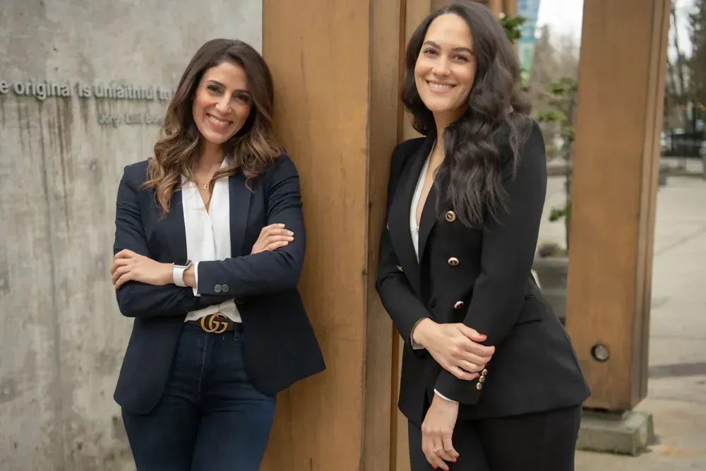 Two women in blazers standing outdoors by a wooden structure. One has arms crossed, smiling. The other leans, smiling.