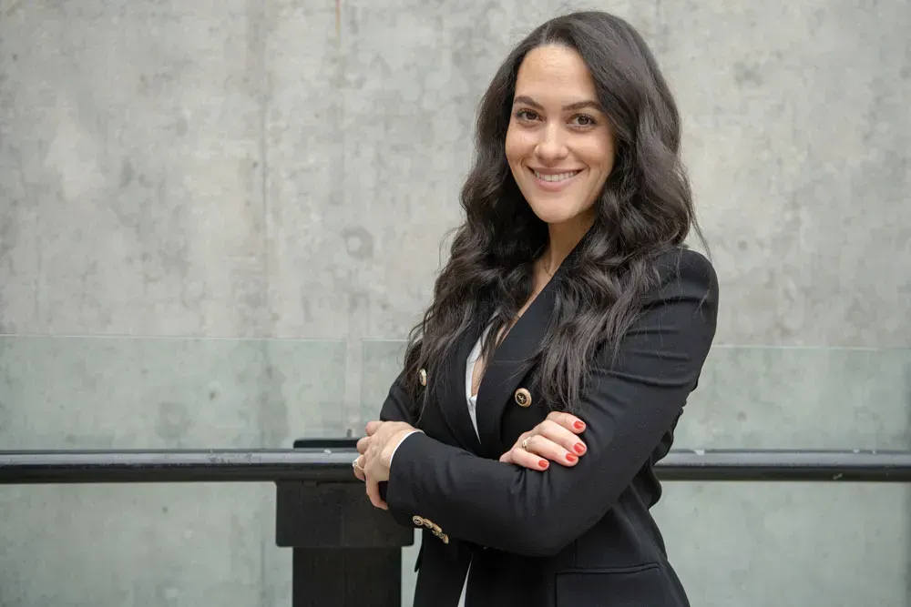 Woman with long dark hair, wearing a black blazer, smiling with arms crossed, leaning on a railing.