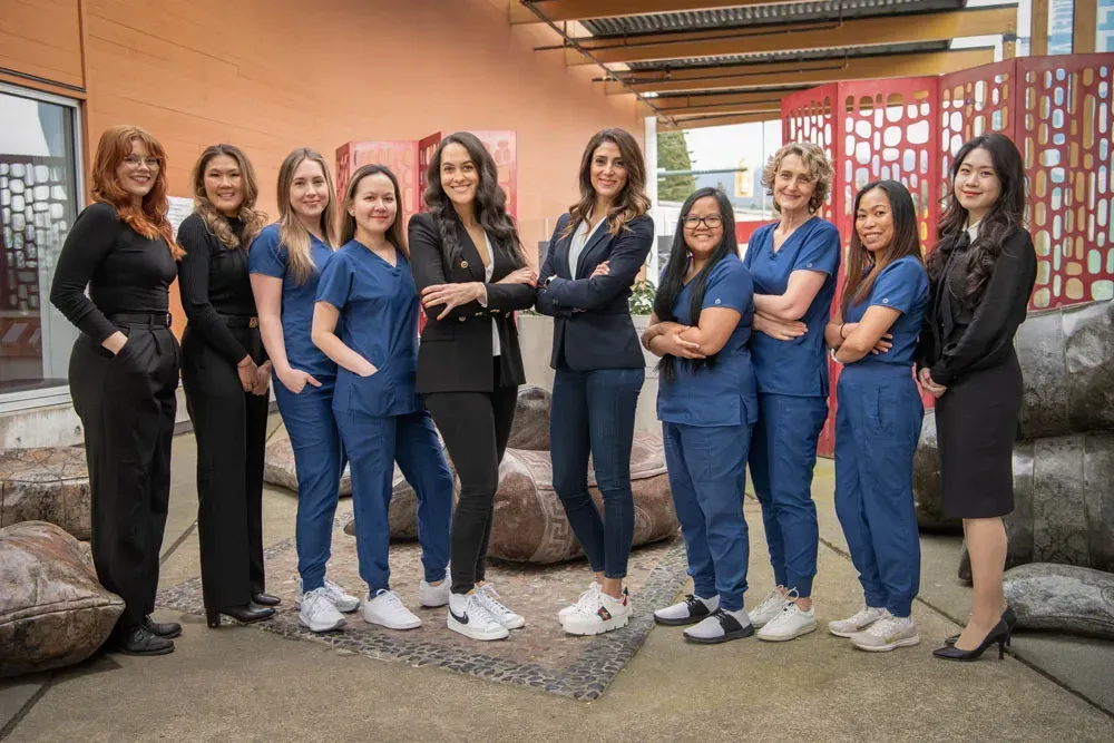 Group of women, mostly smiling, pose outdoors. Some wear scrubs, others business attire. Neutral background.