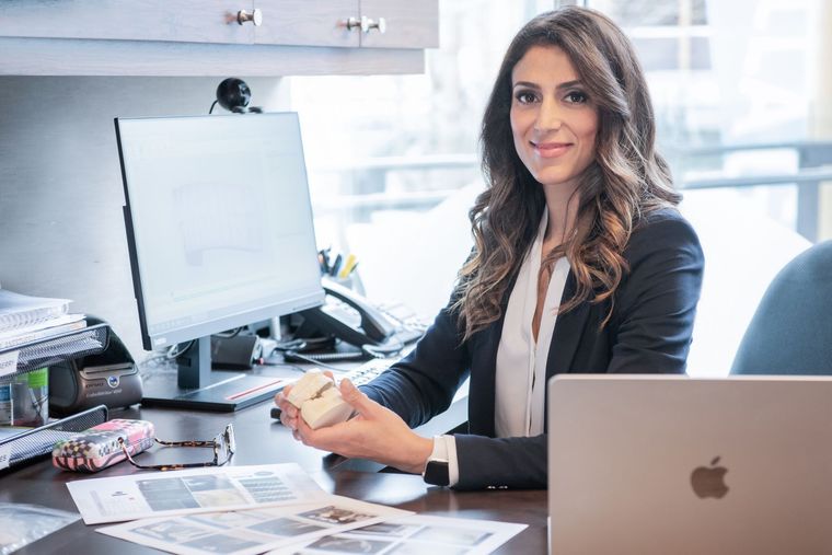 Woman smiling at an office desk with a laptop, desktop monitor, and papers.