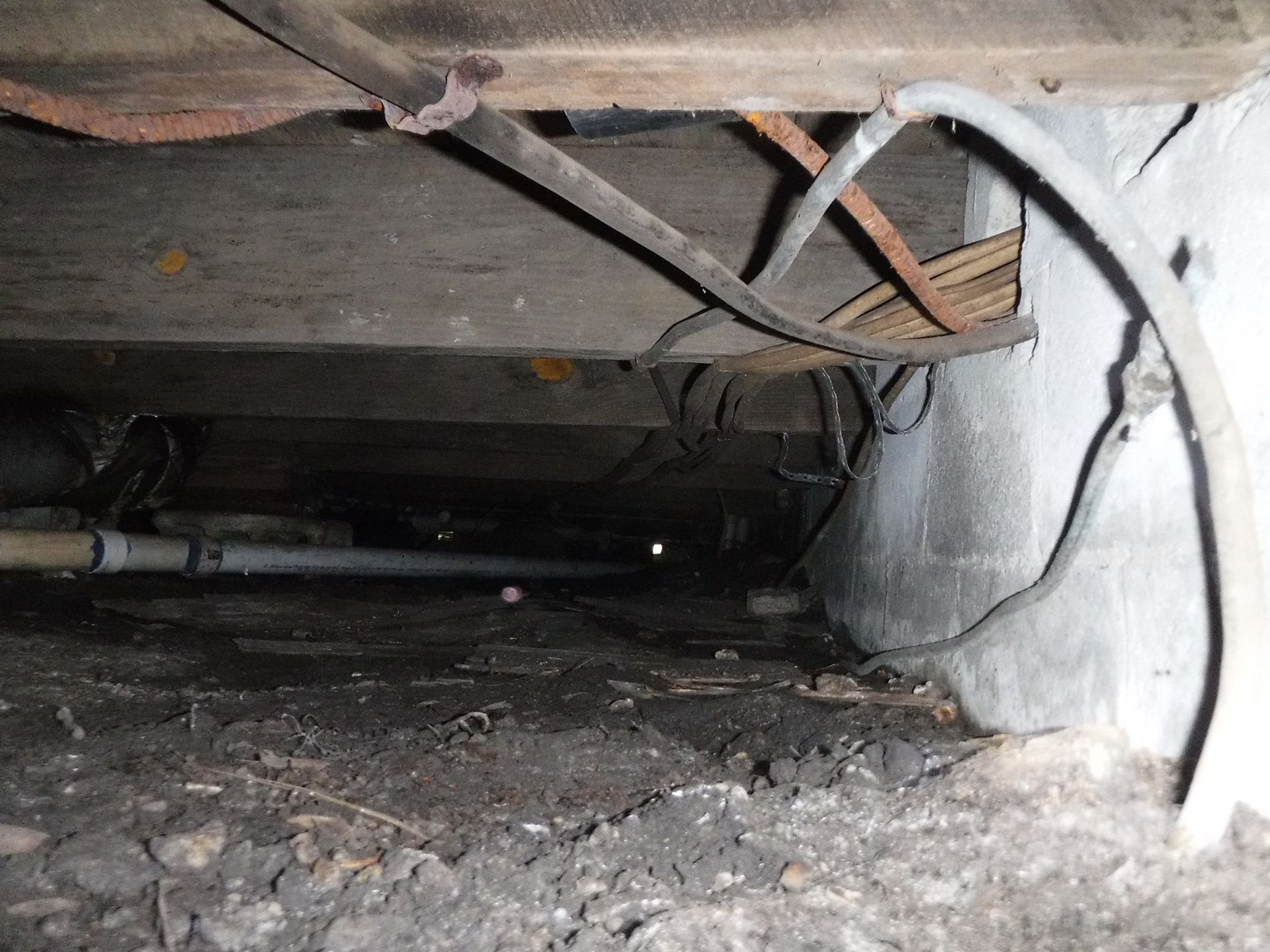 Underneath a structure: dark crawlspace with wires, pipes, and dirt.