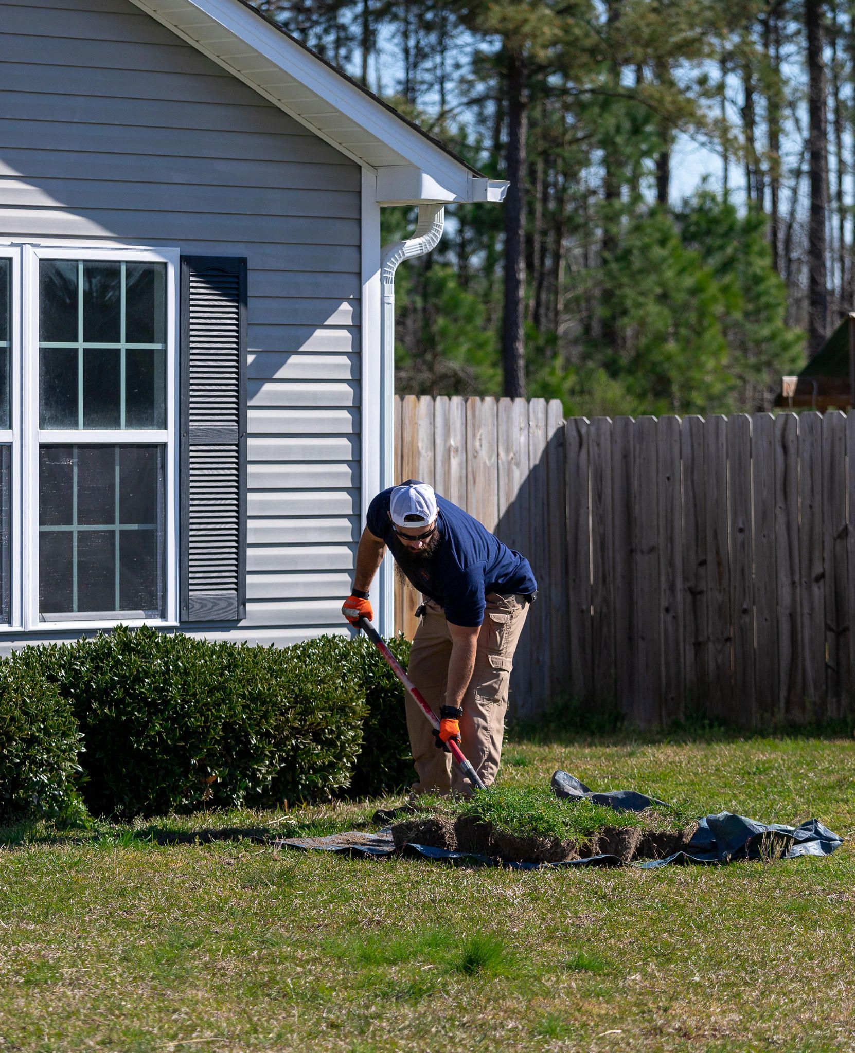 Man using a shovel in a yard near a house and wooden fence.