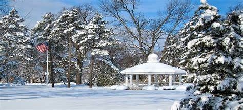 Snow-covered park with a white gazebo, trees, and a flagpole under a clear winter sky