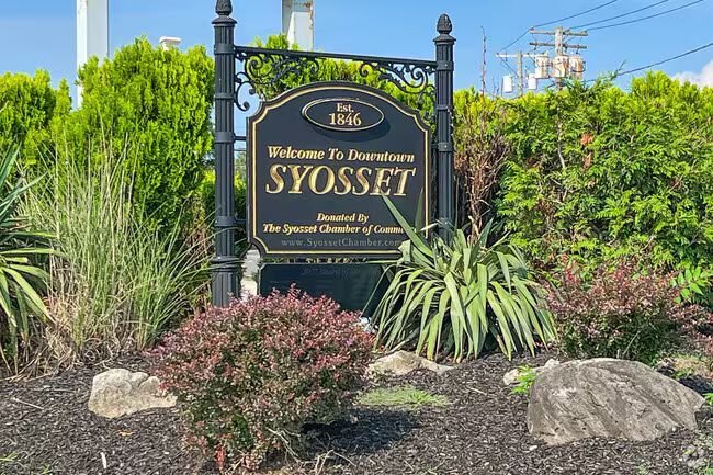 Welcome to Syooset sign surrounded by green shrubs and trees at a landscaped entrance