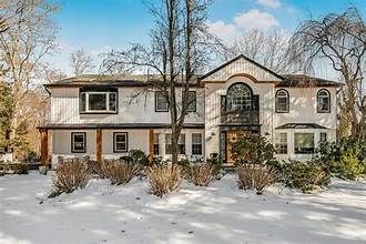 Snow-covered two-story house with beige siding, stone accents, and bare trees in front.