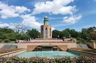 Stone memorial tower with reflecting pool and flowers in a landscaped park under a blue sky